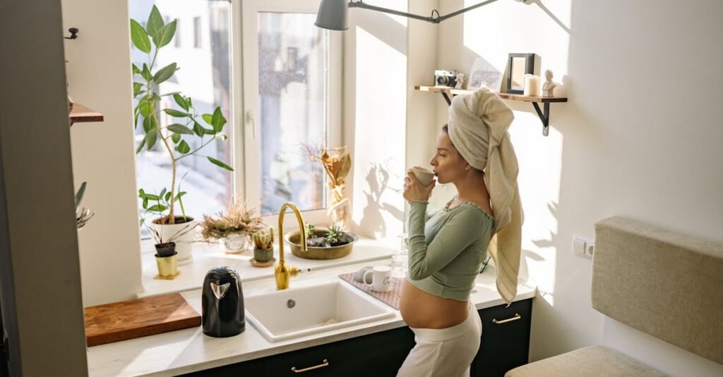 Pregnant woman enjoying a peaceful moment with tea by the window in a sunlit kitchen.
