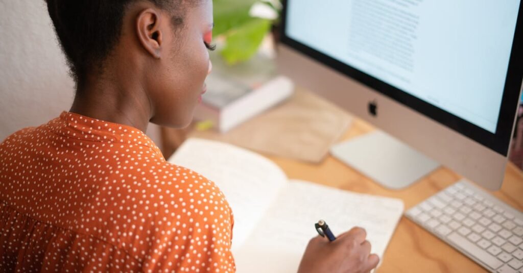 A woman in a polka-dot shirt writes in a notebook at her desk with a computer screen visible.